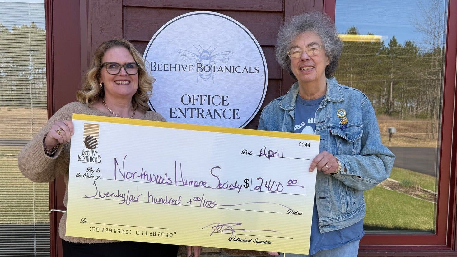 Two women holding a large check in front of a building with 'Beehive Botanicals' sign.