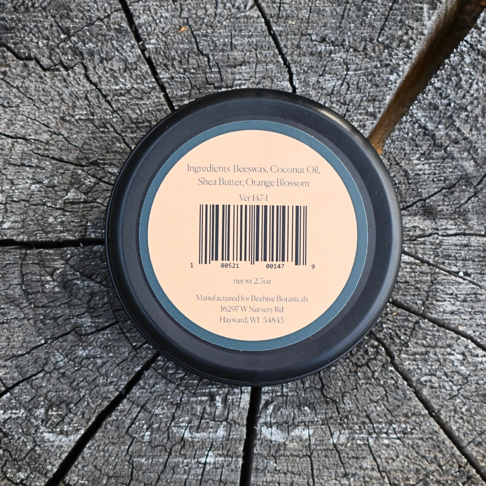 A round black cosmetic tin viewed from above, showing the back label for Beeswax Coconut Oil Shea Butter Orange Blossom balm by Berberine Botanicals, placed on a weathered tree stump.