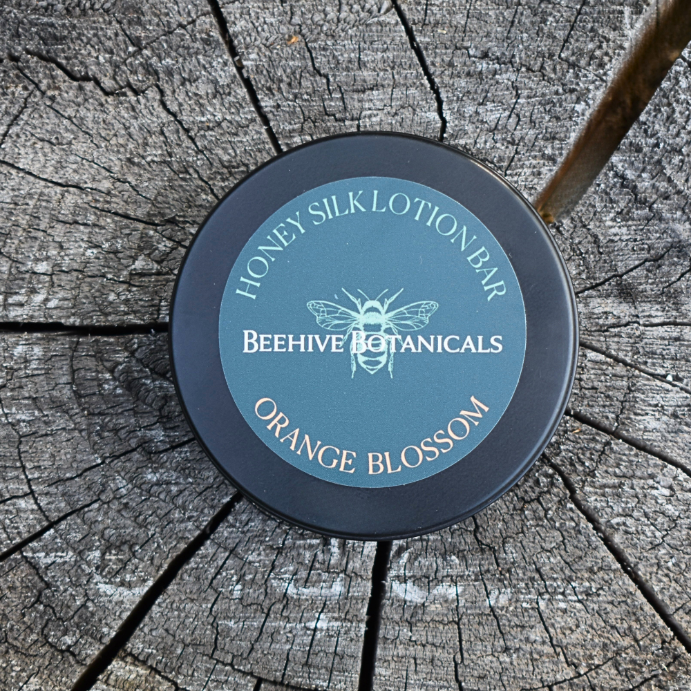 A round black tin of Beehive Botanicals Honey Silk Lotion Bar in Orange Blossom scent, placed on a weathered tree stump. The tin features a dusty blue-green label with a white bee illustration and text.