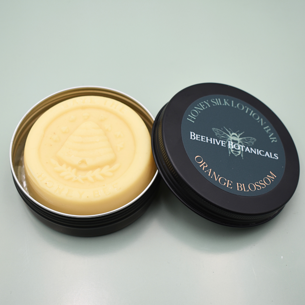 An open round metal tin showing a pale yellow lotion bar, with the black lid beside it. The lid label reads 'HONEY SILK LOTION BAR', 'BEEHIVE BOTANICALS', and 'ORANGE BLOSSOM'.