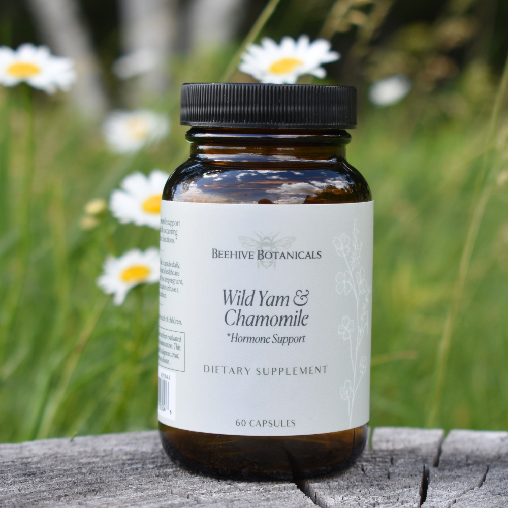 Bottle of Beehive Botanicals Wild Yam & Chamomile supplement on a wooden surface with flowers in the background